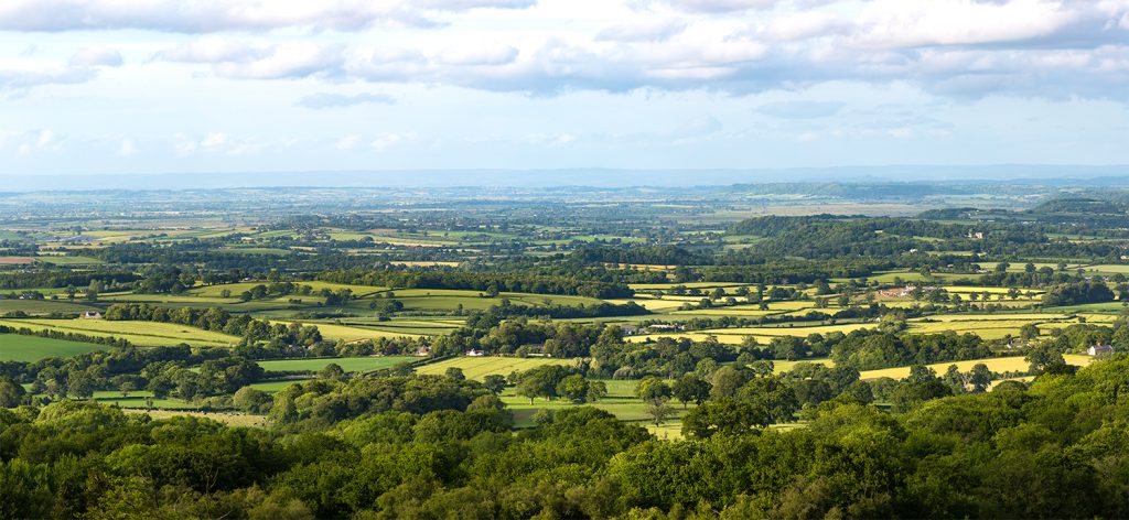 A wide landscape with a patchwork of fields and hedgerows and hills in the backgroun