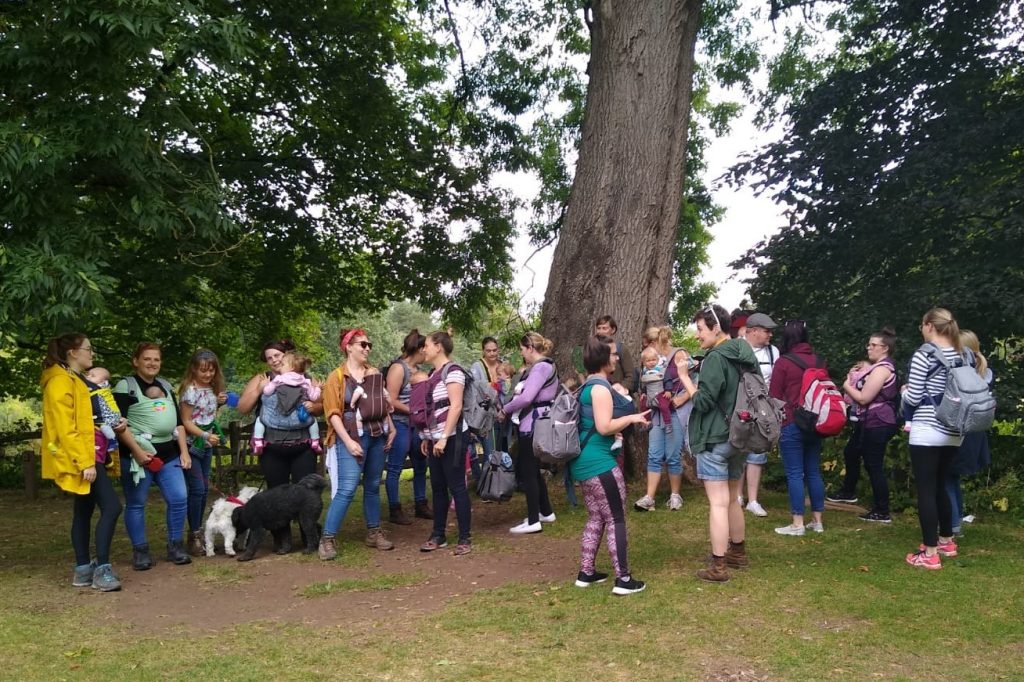 A group of people gathering under a tree with their babies in slings ready for a walk