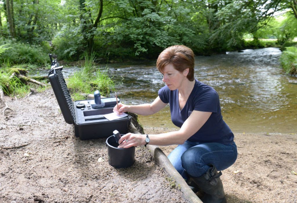 A woman crouched by a river using equipment to measure water quality